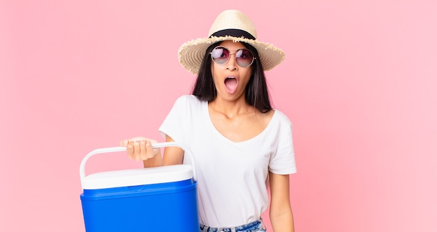 Mulher bonita hispânica parecendo muito chocada ou surpresa com uma geladeira portátil de piquenique