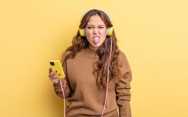 Mulher bonita hispânica se sentindo enojada e irritada e língua para fora fones de ouvido e conceito de smartphone