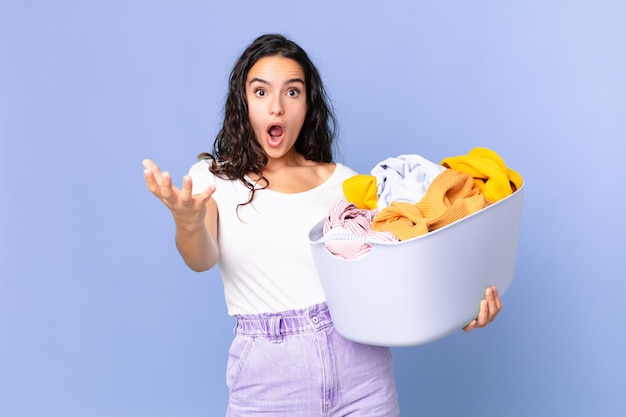 Mulher bonita hispânica se sentindo extremamente chocada e surpresa, segurando uma cesta de roupas para lavar