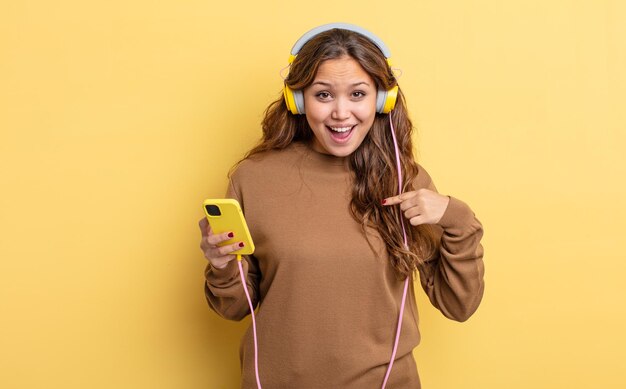 Mulher bonita hispânica se sentindo feliz e apontando para si mesma com um animado. conceito de fones de ouvido e smartphone