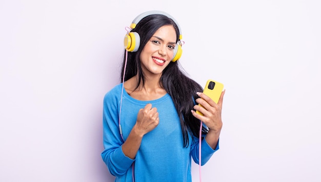 Mulher bonita hispânica se sentindo feliz e enfrentando um desafio ou comemorando. conceito de smartphone e fones de ouvido