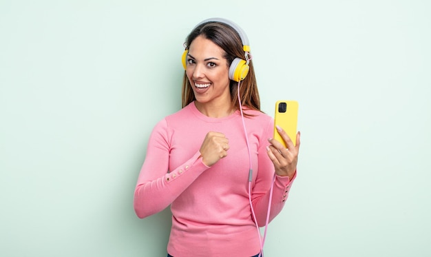 Mulher bonita hispânica se sentindo feliz e enfrentando um desafio ou comemorando. conceito de smartphone e fones de ouvido