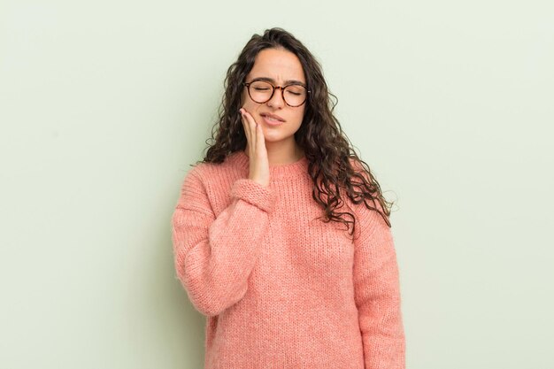 Mulher bonita hispânica segurando a bochecha e sofrendo dor de dente dolorosa, sentindo-se doente, miserável e infeliz, procurando um dentista