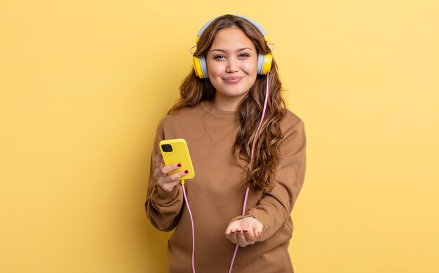 Mulher bonita hispânica sorrindo alegremente com amigável e oferecendo e mostrando um conceito de fones de ouvido e smartphone