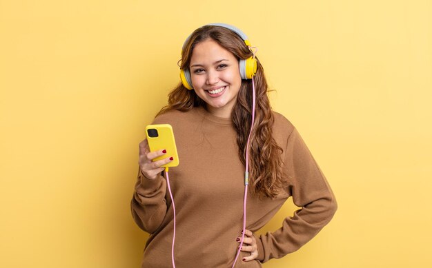 Mulher bonita hispânica, sorrindo alegremente com uma mão no quadril e confiante. conceito de fones de ouvido e smartphone