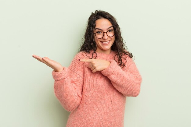 Mulher bonita hispânica sorrindo alegremente e apontando para copiar o espaço na palma da mão mostrando ou anunciando um objeto