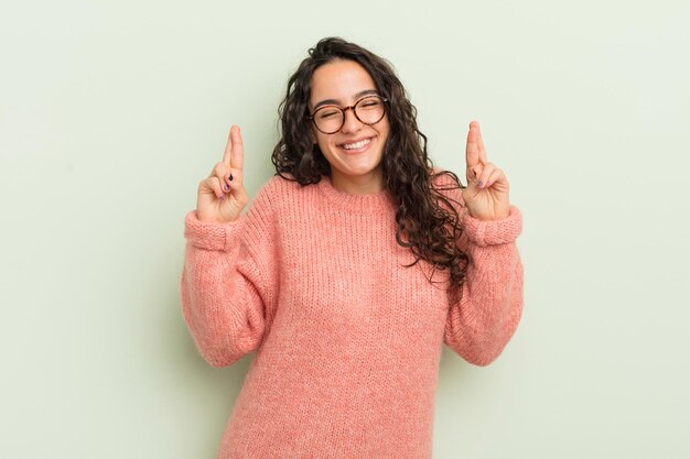 Mulher bonita hispânica sorrindo e cruzando ansiosamente os dois dedos sentindo-se preocupada e desejando ou esperando boa sorte