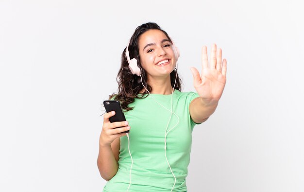 Mulher bonita hispânica sorrindo e parecendo amigável, mostrando o número cinco com fones de ouvido e um smartphone