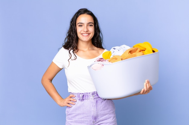 Mulher bonita hispânica sorrindo feliz com uma mão no quadril e confiante, segurando uma cesta para lavar roupas