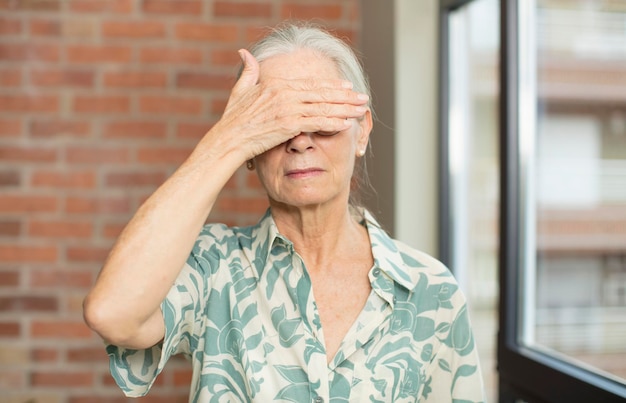 Mulher bonita idosa cobrindo os olhos com uma mão, sentindo-se assustada ou ansiosa, imaginando ou esperando cegamente por uma surpresa