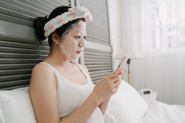 mulher bonita japonesa asiática com máscara facial usando telefone celular no quarto de manhã. jovem encantadora concentrada olhando para smartphone enquanto estava deitado na cama esperando o tempo de cuidados com a pele de beleza em casa.