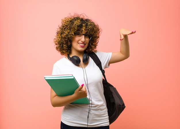 Mulher bonita jovem estudante sorrindo, sentindo-se feliz, positivo e satisfeito, segurando ou mostrando objeto ou conceito no espaço da cópia contra parede rosa