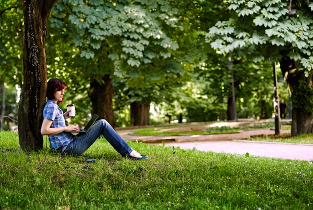 Mulher bonita jovem freelancer usando laptop sentado debaixo da árvore no parque.