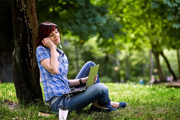 Mulher bonita jovem freelancer usando laptop sentado debaixo da árvore no parque.