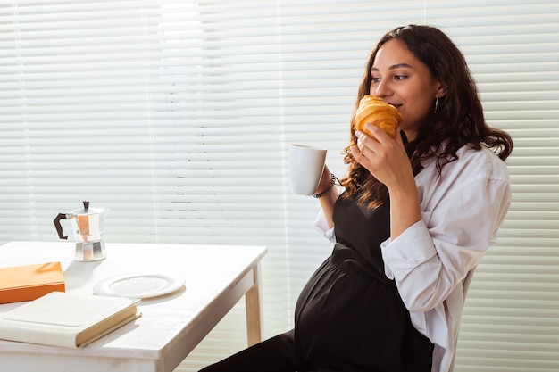 Mulher bonita jovem grávida feliz comendo croissant durante o conceito de café da manhã agradável