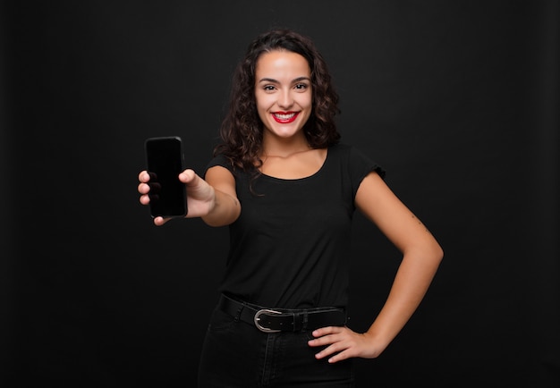 Mulher bonita jovem sorrindo alegremente com uma mão no quadril e atitude confiante, positiva, orgulhosa e amigável, segurando um smartphone