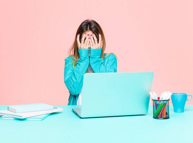 Foto mulher bonita jovem trabalhando com um laptop sentindo medo ou vergonha, espiando ou espionando com os olhos semicobertos com as mãos