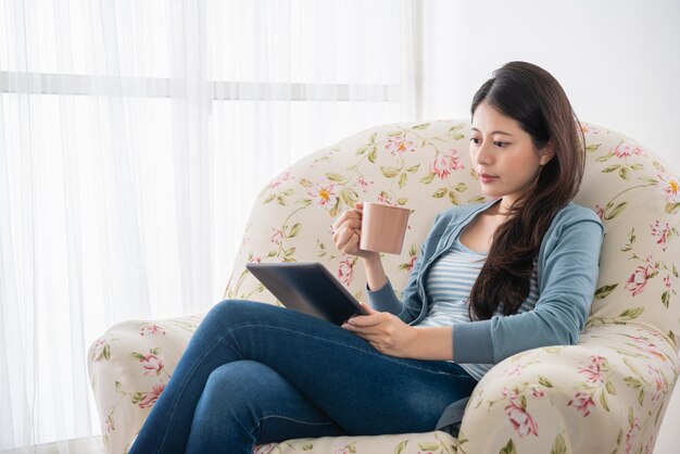 mulher bonita lendo tablet e segurando um policial de café sentado no sofá com um olhar relaxante.