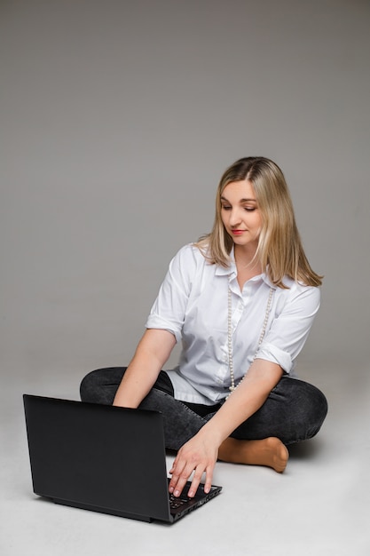 Mulher bonita loira com camisa branca, jeans escuros e miçangas no pescoço digitando no laptop sentado com as pernas cruzadas no chão. isole em cinza.