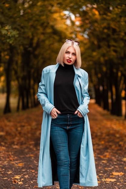 Mulher bonita loira com lábios vermelhos na moda casaco azul, jeans e suéter preto passeios no parque outono. Beleza e estilo feminino de outono