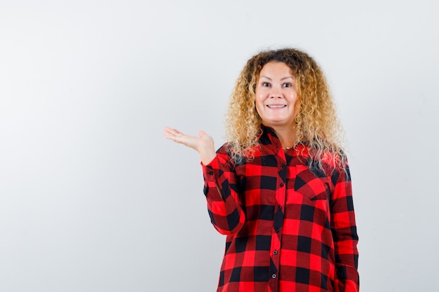 Mulher bonita loira de camisa xadrez fingindo segurar algo e olhando alegre, vista frontal.