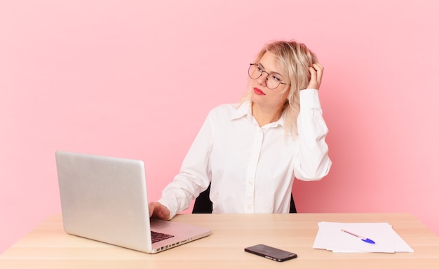 Mulher bonita loira jovem bonita sentindo-se perplexo e confuso, coçando a cabeça. conceito de mesa de trabalho