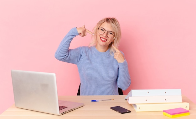 Mulher bonita loira jovem bonita sorrindo com confiança, apontando para o próprio sorriso largo. conceito de mesa de trabalho