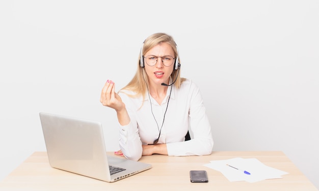 Mulher bonita loira jovem loira fazendo um gesto de capice ou dinheiro, dizendo para você pagar e trabalhando com um laptop