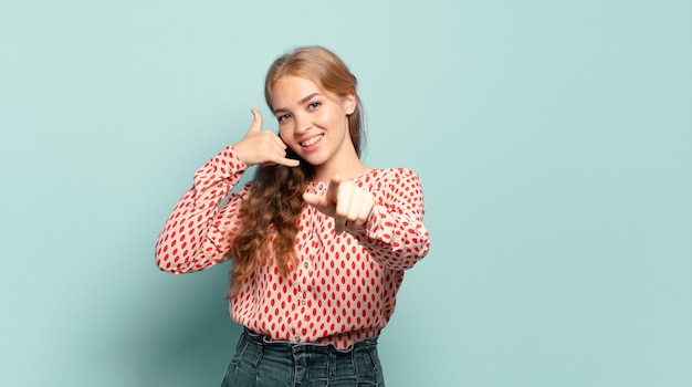 Mulher bonita loira sorrindo alegremente e apontando para a frente enquanto faz um gesto para ligar para você, falando ao telefone