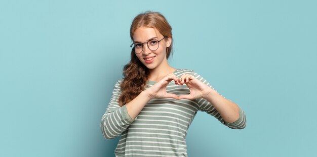 Mulher bonita loira sorrindo e se sentindo feliz, fofa, romântica e apaixonada, fazendo formato de coração com as duas mãos