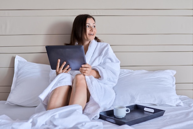 Mulher bonita madura descansando no hotel. Fêmea em roupão branco com xícara de café gostando de assistir tablet digital, sentado na cama