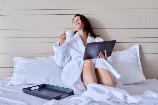 Mulher bonita madura descansando no hotel Mulher em roupão branco com xícara de café gostando de assistir tablet digital sentado na cama