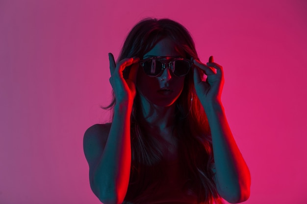 Mulher bonita moderna jovem hippie no topo da moda coloca óculos de sol elegantes em uma sala com uma luz vermelha incrível. Modelo de moda de menina retrato com lábios sensuais dentro de casa com cor neon em estilo discoteca.