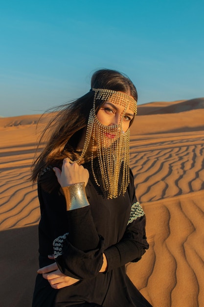 Mulher bonita na máscara Burqa brilhante entre o deserto de Dubai ao pôr do sol estilo árabe