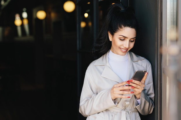 Mulher bonita na moda com cabelo escuro amarrado em rabo de cavalo vestido com casaco branco elegante segurando o celular olhando para a tela com expressão deliciosa jovem modelo bonito se comunicando online