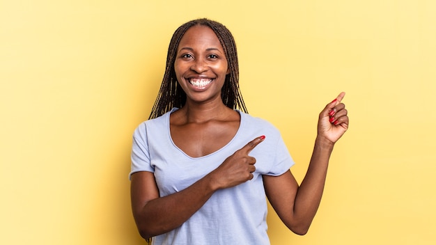 Mulher bonita negra afro sorrindo alegremente e apontando para o lado e para cima com as duas mãos mostrando o objeto no espaço da cópia