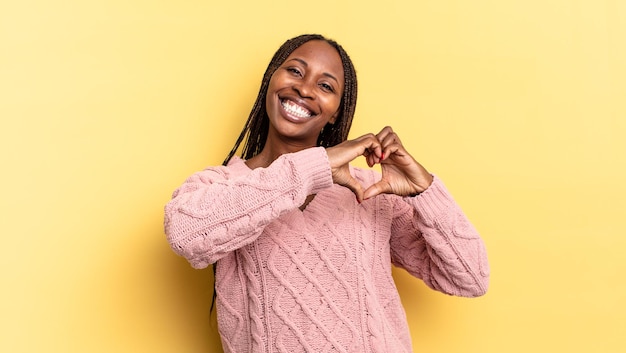 Mulher bonita negra afro sorrindo e se sentindo feliz, fofa, romântica e apaixonada, fazendo formato de coração com as duas mãos