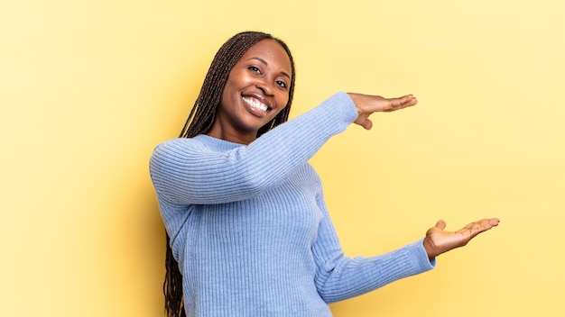Mulher bonita negra afro sorrindo, sentindo-se feliz, positiva e satisfeita, segurando ou mostrando um objeto ou conceito no espaço da cópia
