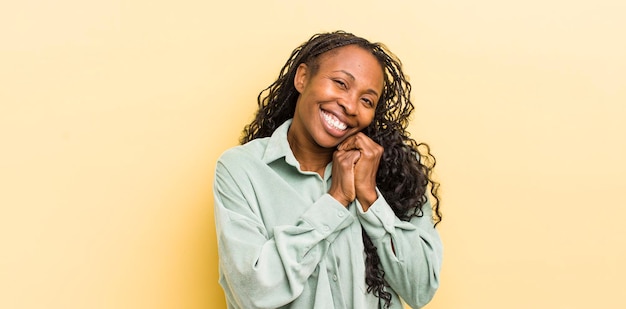 Mulher bonita negra se sentindo apaixonada e parecendo fofa adorável e feliz sorrindo romanticamente com as mãos ao lado do rosto