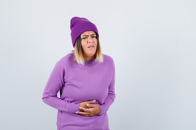 Mulher bonita no suéter, gorro sofrendo de dor de estômago e parecendo doente, vista frontal.