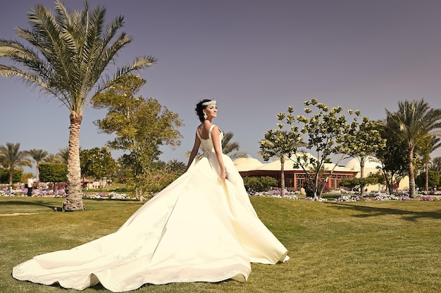 Mulher bonita no vestido de noiva na cor branca, a menina tem maquiagem da moda e cabelo em pé ao ar livre, verão ensolarado em fundo natural, noiva em celebração do feriado