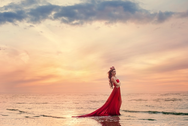 Mulher bonita no vestido vermelho esvoaçante na onda do mar ao pôr do sol.
