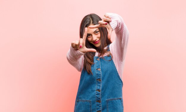 Mulher bonita nova que sente feliz, amigável e positiva, sorrindo e fazendo um retrato ou uma moldura com as mãos contra a parede rosa.