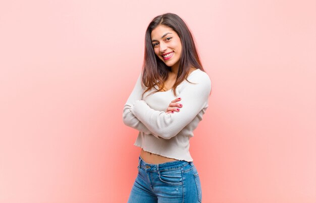 Mulher bonita nova que sorri com braços cruzados e uma expressão feliz, segura, satisfeita, vista lateral contra a parede cor-de-rosa.