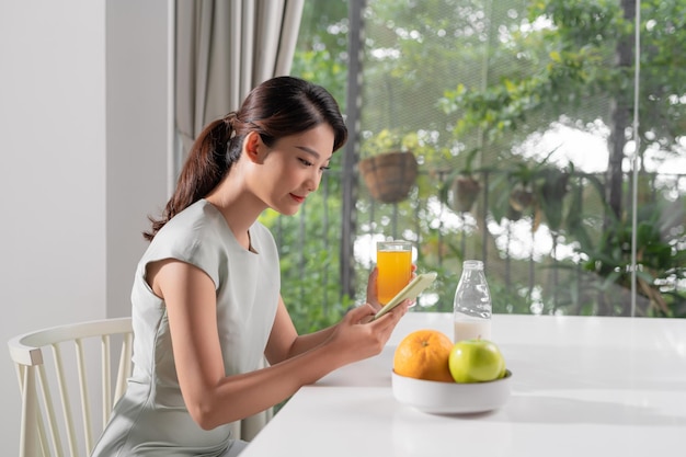 Mulher bonita olhando para o celular e segurando o copo de suco de laranja enquanto toma café da manhã