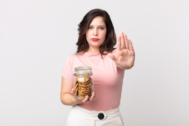 Mulher bonita olhando séria mostrando a palma da mão aberta fazendo gesto de pare e segurando uma garrafa de vidro de biscoitos