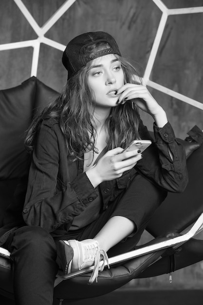 Mulher bonita ou linda garota sexy com cabelo comprido em boné preto e tênis sentado na cadeira geométrica segurando celular ou celular em fundo cinza
