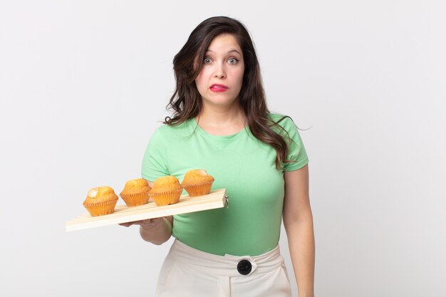 Mulher bonita parecendo perplexa e confusa segurando uma bandeja de muffins