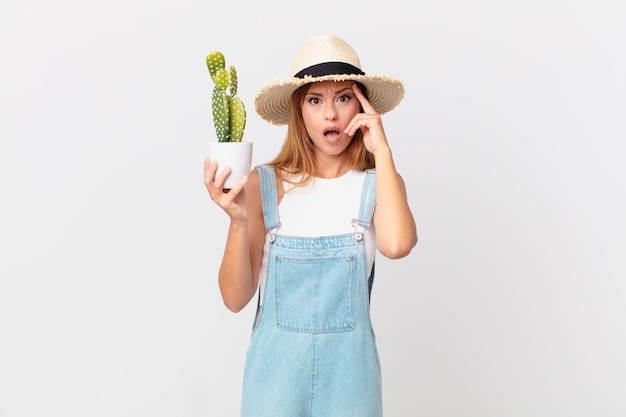 Mulher bonita parecendo surpresa, percebendo um novo pensamento, ideia ou conceito e segurando uma planta decorativa de cacto