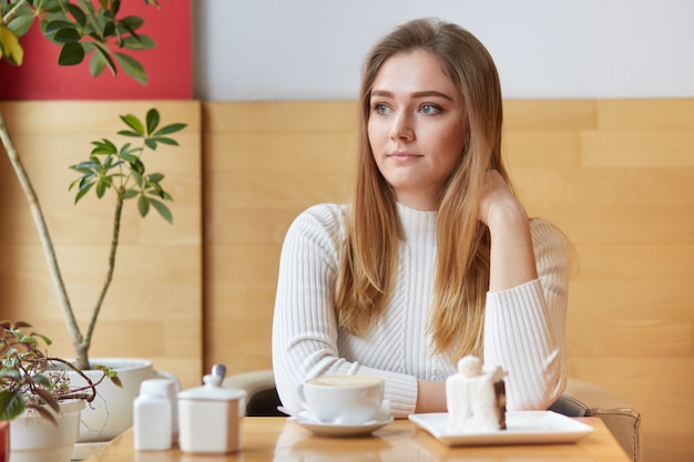 Mulher bonita pensativa focada pensativamente de lado, bebe café expresso e come bolo delicioso na cafeteria